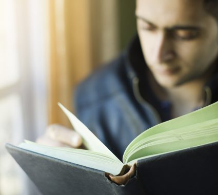 Indoor day time image of a defocused young man studying at home sitting near window, the focus is on the book he is holding and turning the page while looking down at the book. One person, waist up, horizontal composition with selective focus and copy space.